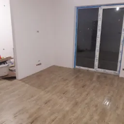 Wnętrze pokoju z nową podłogą imitującą drewno, świeżo pomalowane ściany na biało, duże okno przesuwne z niebieską folią ochronną na ramie, widoczne gniazdka elektryczne i fragment pomieszczenia...