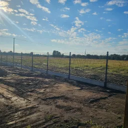 Długi, prosty odcinek ogrodzenia panelowego z podmurówką betonową w trakcie montażu na nierównym, przygotowanym terenie. Widoczne ślady po maszynach budowlanych na ziemi, błękitne niebo z chmurami...