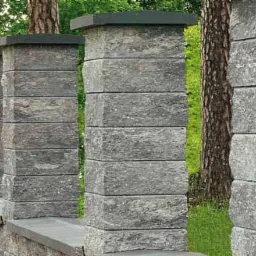 Szary, betonowy płot modułowy z ciemnymi nakrywami, widok z boku, drzewo wkomponowane w ogrodzenie.