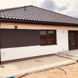 Nowoczesny dom jednorodzinny z elewacją w kolorach bieli i grafitu, z ciemną dachówką i drewnianymi żaluzjami zewnętrznymi, widok z zewnątrz w trakcie prac wykończeniowych.