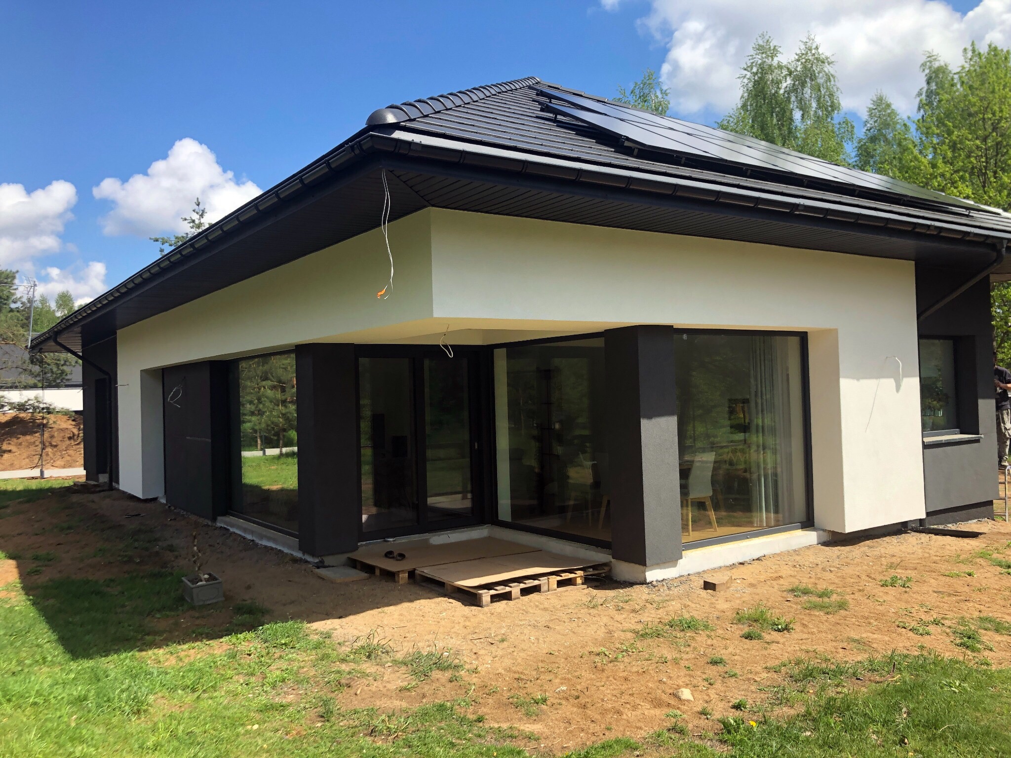 Nowoczesny dom jednorodzinny z płaskim dachem i panelami słonecznymi, wykończony w kolorach bieli i grafitu, z dużymi oknami, widoczne przygotowania do zagospodarowania terenu wokół budynku.