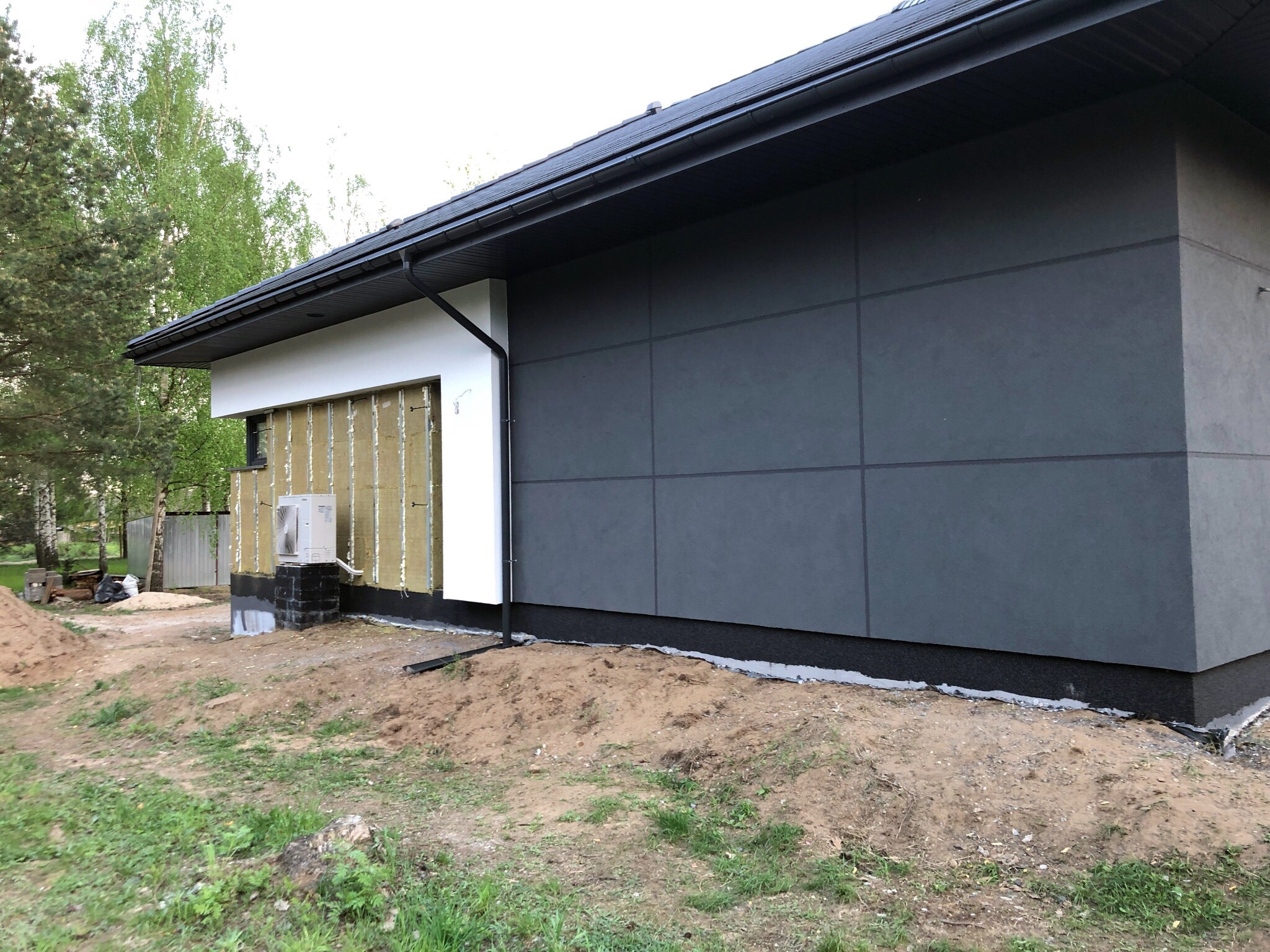 Dom w trakcie prac elewacyjnych: szara elewacja w panelach, fragment ocieplenia z wełny mineralnej widoczny obok klimatyzatora, czarny dach i rynna.