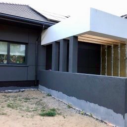 Szary dom jednorodzinny z fragmentem elewacji pokrytej panelami i otynkowanym murem, widoczne elementy konstrukcji dachu i izolacji w części dobudowanej.