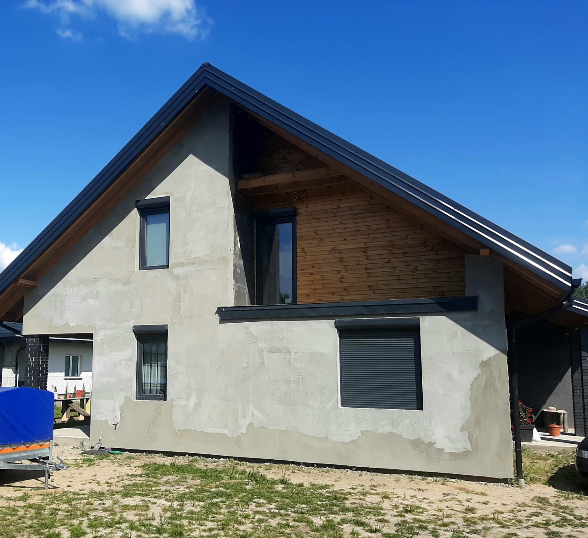 Dom w trakcie nakładania tynku zewnętrznego, widoczne fragmenty surowego tynku, okna z roletami zewnętrznymi i drewniane wykończenie pod dachem.