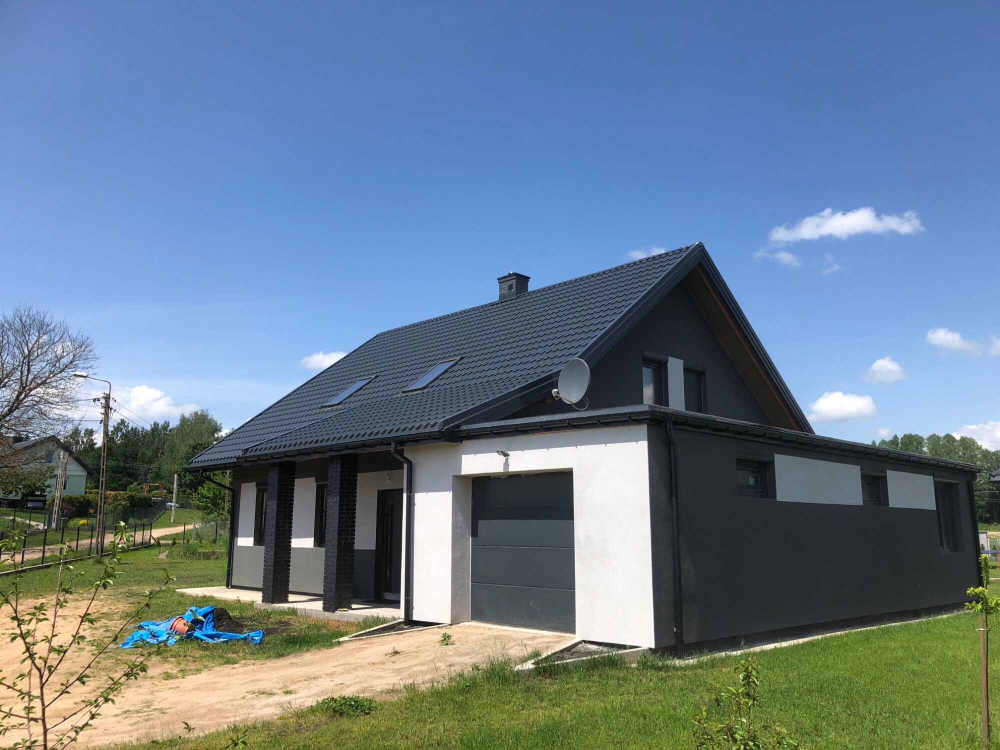 Nowoczesny dom jednorodzinny z grafitową elewacją i białymi akcentami, widok z zewnątrz w słoneczny dzień, na trawniku widoczna niebieska plandeka.