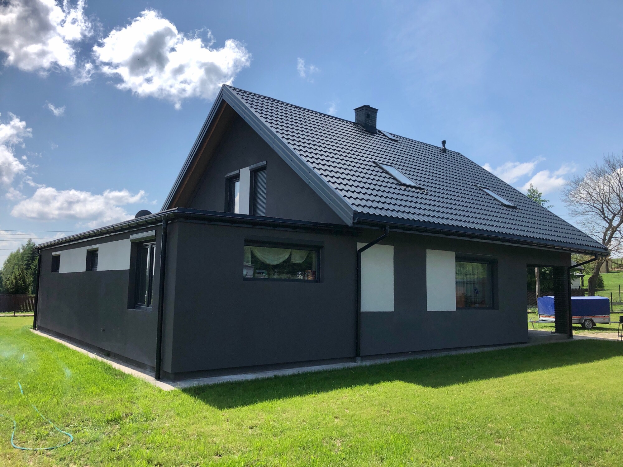 Nowoczesny dom jednorodzinny z ciemną elewacją i grafitową dachówką, widok z trawnika w słoneczny dzień.