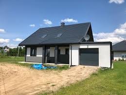 Nowoczesny dom jednorodzinny z szarą dachówką i elewacją w kolorach bieli i grafitu, widoczny garaż z szarą bramą, teren wokół domu w trakcie prac wykończeniowych, na piasku leży zwinięta niebieska...