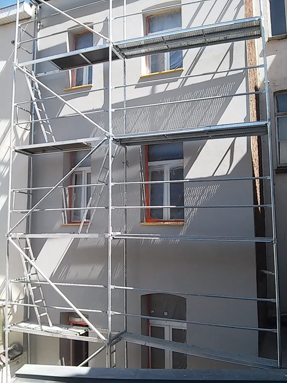 Elewacja budynku w trakcie prac tynkarskich, z widocznymi rusztowaniami i oknami zabezpieczonymi folią.