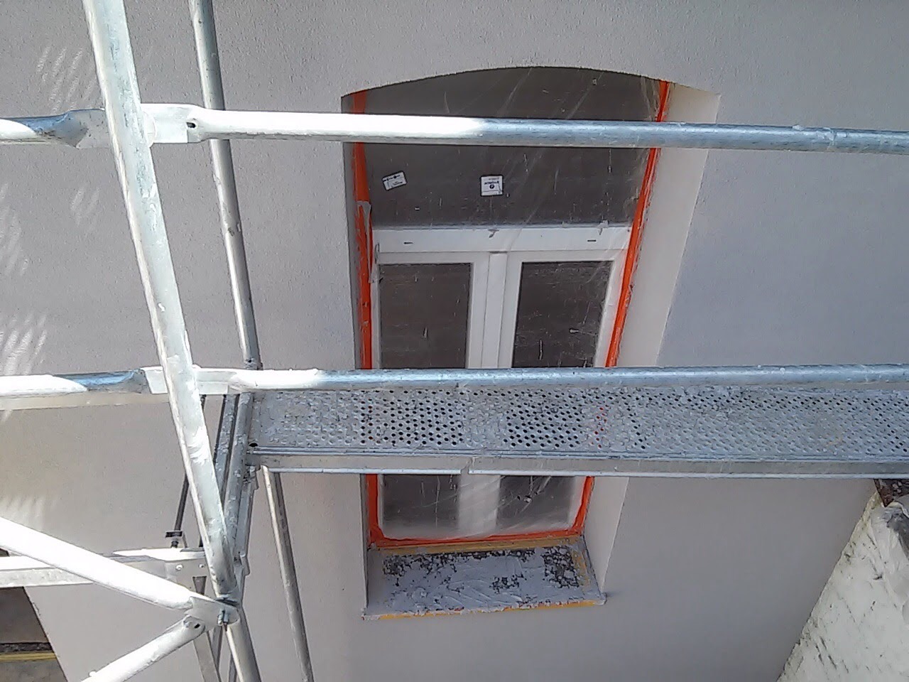 Okno dachowe zabezpieczone folią malarską podczas prac elewacyjnych, widok z rusztowania, świeżo nałożona szara warstwa tynku.