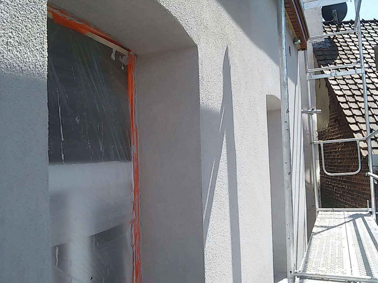 Świeżo otynkowana elewacja budynku z zabezpieczonym oknem folią i taśmą malarską, widoczny fragment rusztowania i cień na ścianie.