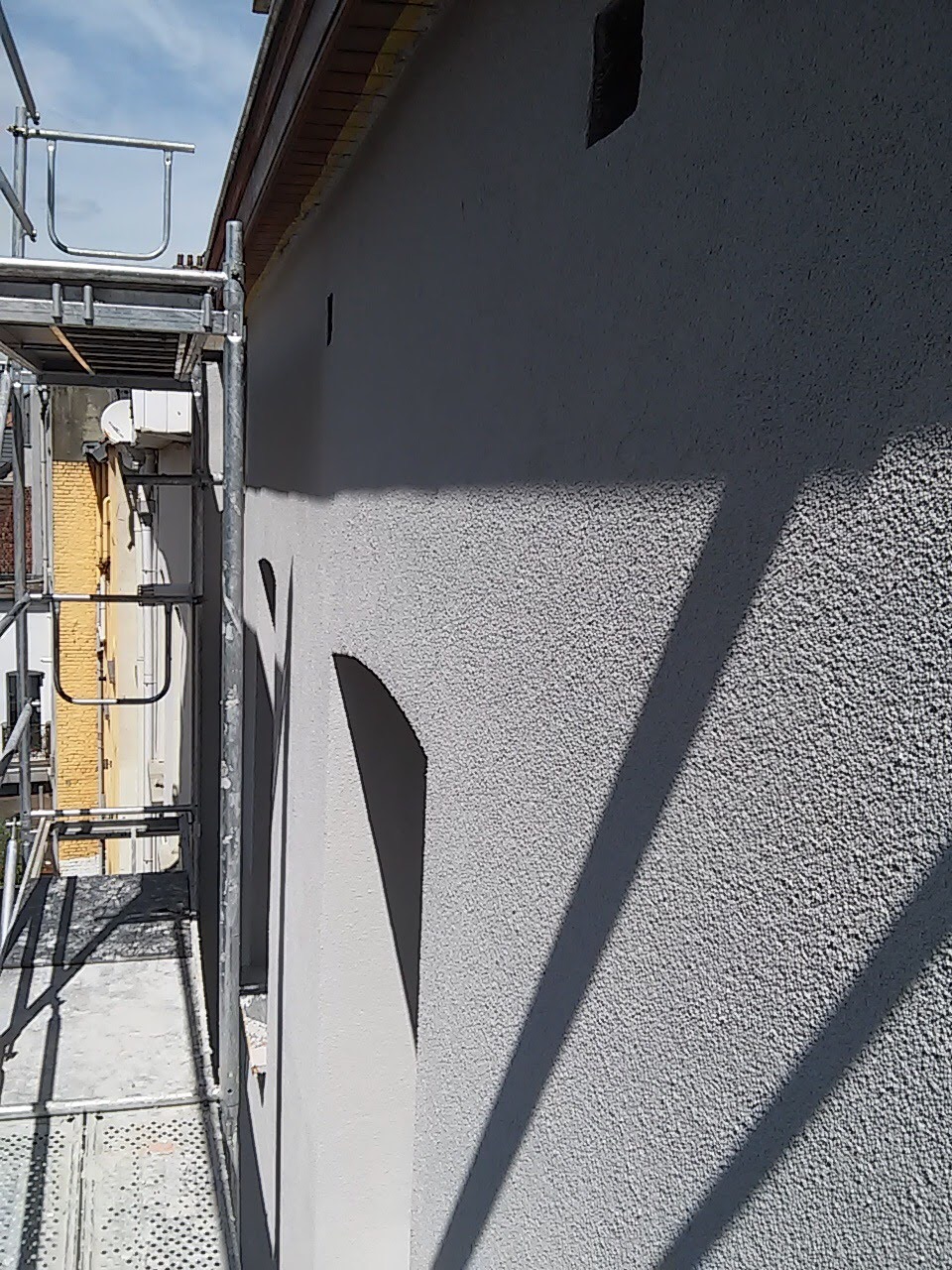 Świeżo otynkowana, jasnoszara elewacja budynku z widocznym rusztowaniem i cieniem, ukazująca fakturę tynku baranka.