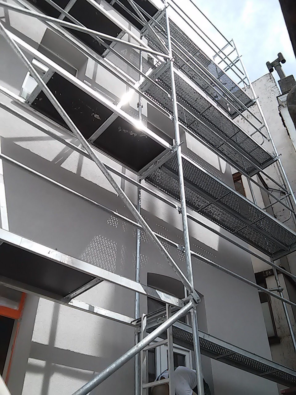 Świeżo pomalowana elewacja budynku z ustawionymi metalowymi rusztowaniami, widoczne refleksy słońca na ścianie i elementach rusztowania.
