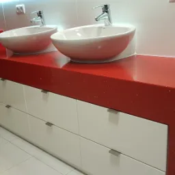 Łazienka z dwoma okrągłymi umywalkami nablatowymi na czerwonym blacie z białymi szafkami z szufladami i srebrnymi uchwytami.