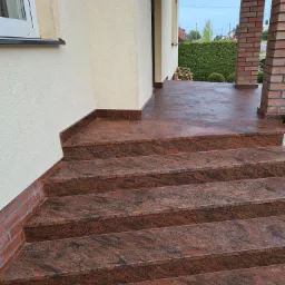 Zewnętrzne schody wejściowe wyłożone czerwono-brązowym kamieniem z wyraźnym użyleniem, prowadzące na taras z ceglanymi filarami.