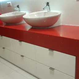Czerwony, błyszczący blat z konglomeratu z dwiema owalnymi umywalkami nablatowymi i chromowanymi bateriami, zamontowany na białych szafkach z szufladami, w nowoczesnej łazience.