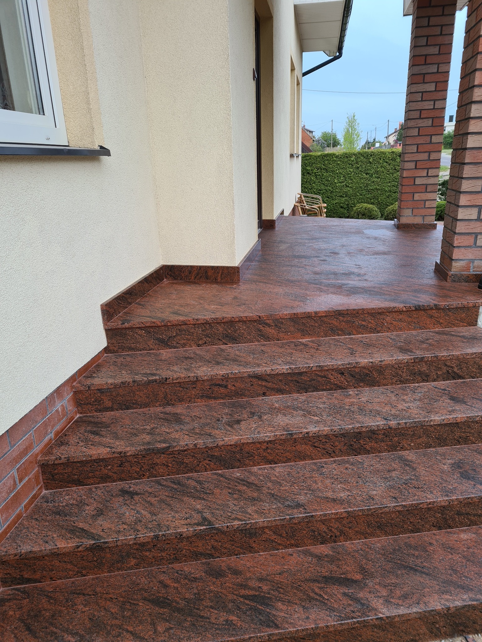 Zewnętrzne schody wejściowe wyłożone czerwono-brązowym kamieniem z wyraźnym użyleniem, prowadzące na taras z ceglanymi filarami.