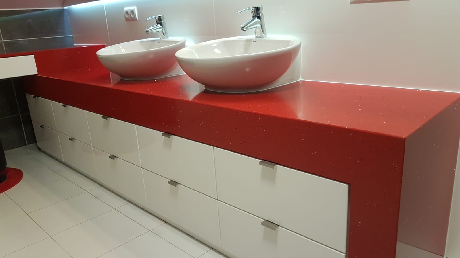 Czerwony, błyszczący blat z konglomeratu z dwiema owalnymi umywalkami nablatowymi i chromowanymi bateriami, zamontowany na białych szafkach z szufladami, w nowoczesnej łazience.