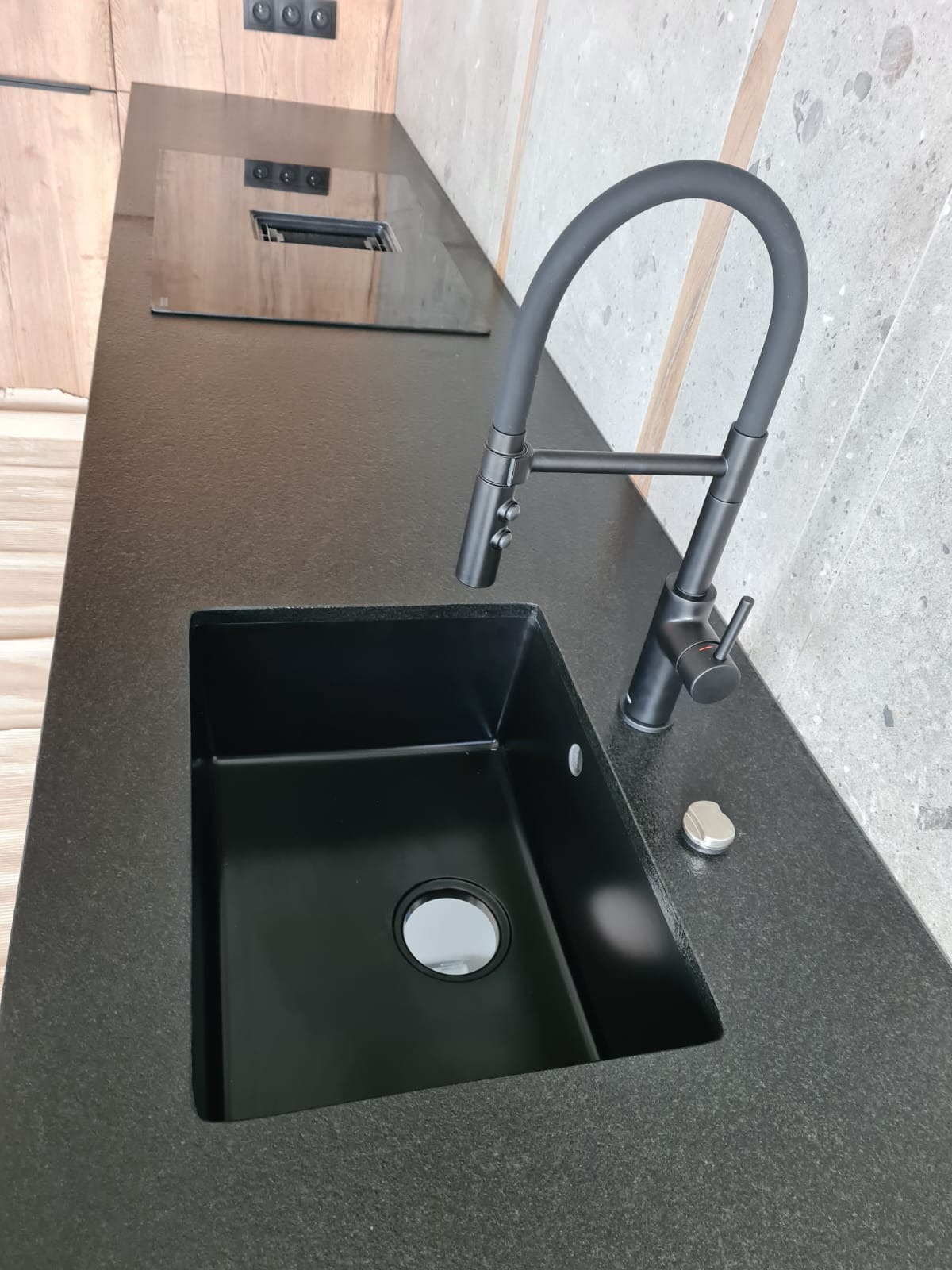Czarny, matowy zlew z baterią o giętkiej wylewce, wkomponowany w ciemny, nakrapiany blat. Widoczny dozownik na płyn. Tło stanowi drewniana zabudowa i szara ściana.