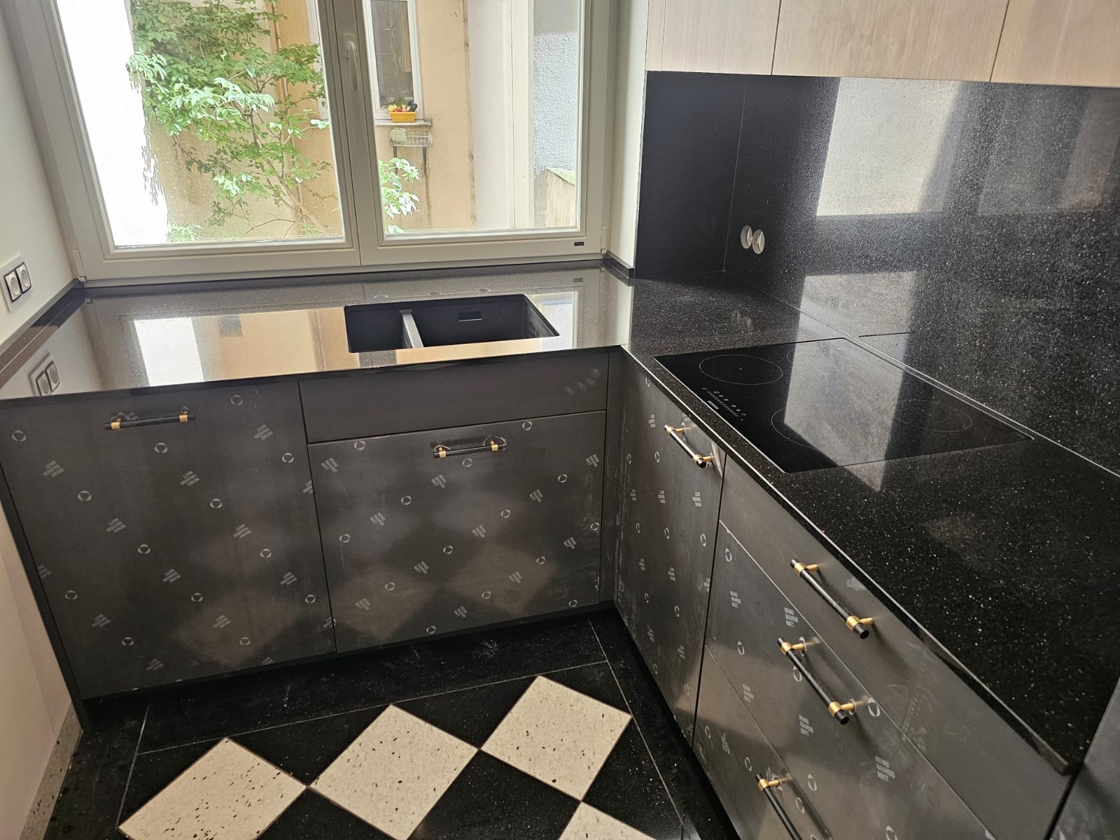 Czarny, błyszczący blat kuchenny z wbudowanym zlewem i płytą indukcyjną, wzorzyste fronty szafek, podłoga w czarno-białą szachownicę, widok na zieleń za oknem.