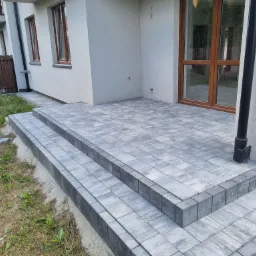 Nowy taras z szarej kostki brukowej prowadzący do wejścia domu, z widocznymi schodami i fragmentami niezakończonych prac w postaci stosu kostki obok.