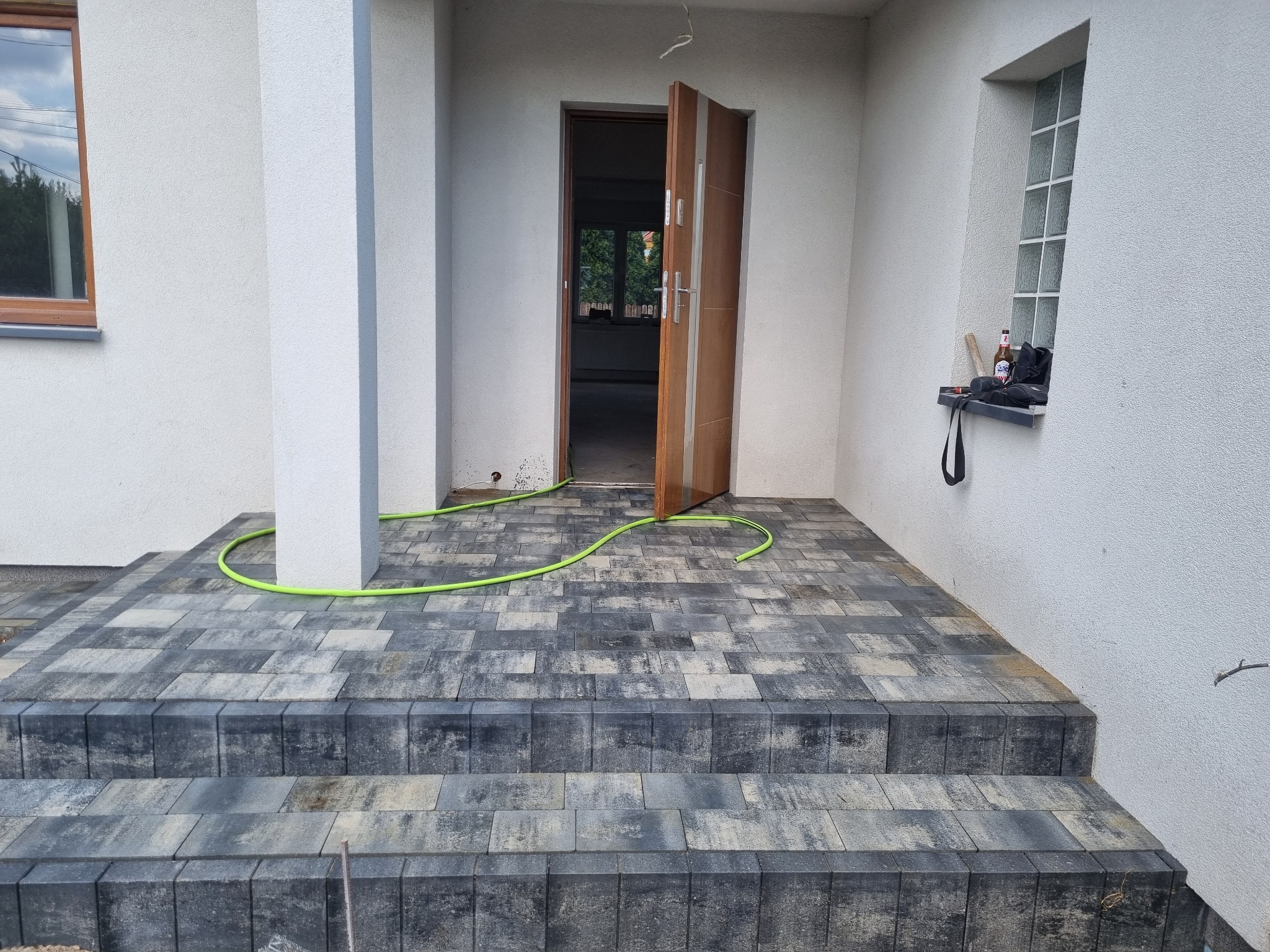Nowo ułożona, szara kostka brukowa na schodach wejściowych do domu, z widocznym fragmentem zielonego węża ogrodowego i otwartymi drzwiami wejściowymi.