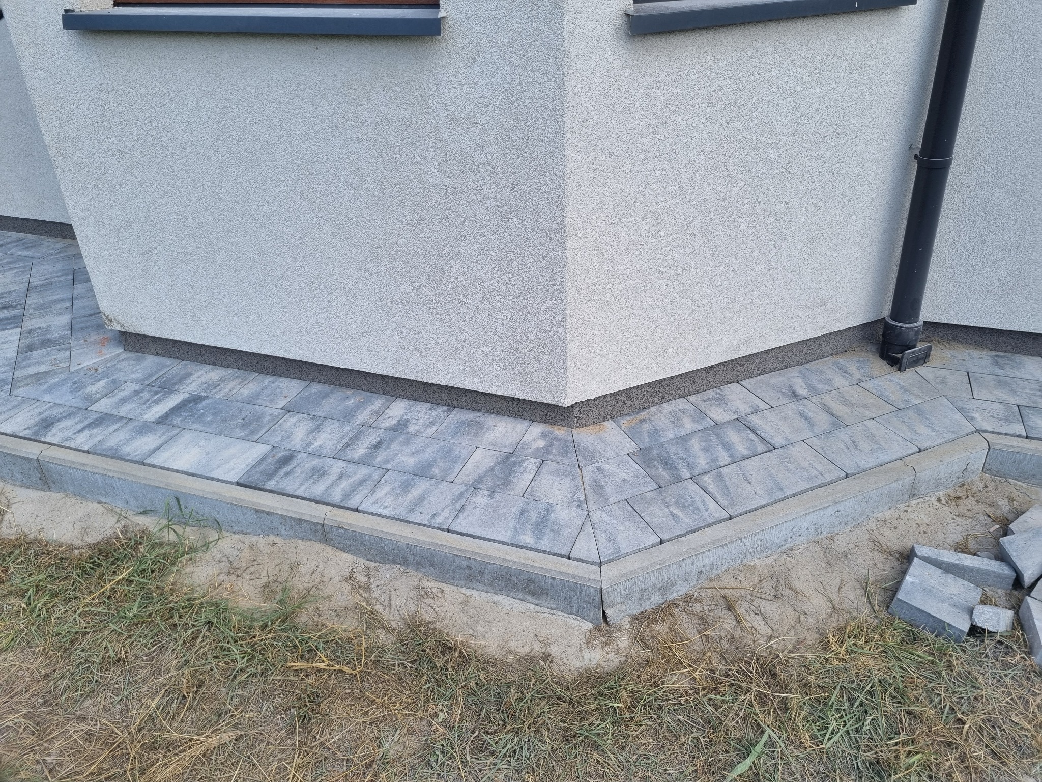 Wykończony narożnik domu z ułożoną szarą kostką brukową, widoczny fragment elewacji z oknem i rurą spustową, oraz niezabudowany fragment z piaskiem i trawą.