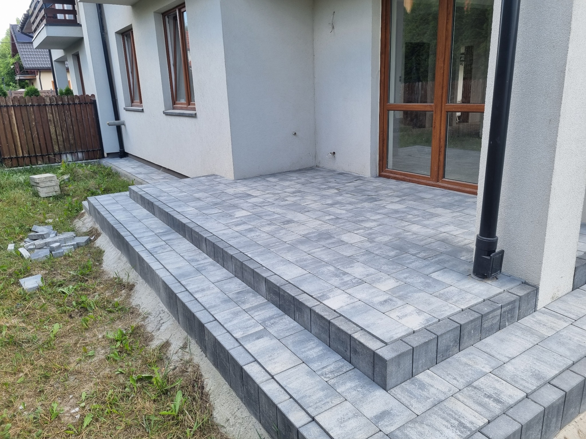 Nowy taras z szarej kostki brukowej prowadzący do wejścia domu, z widocznymi schodami i fragmentami niezakończonych prac w postaci stosu kostki obok.