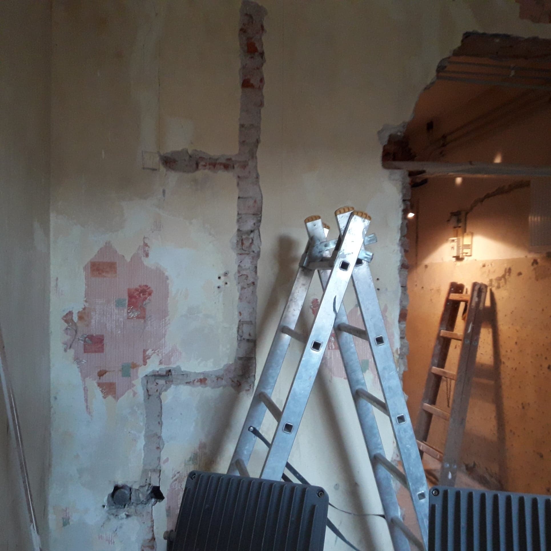 Demontaż starej instalacji elektrycznej w mieszkaniu: Wykute bruzdy w ścianie z resztkami tapety, odsłonięte przewody, aluminiowa drabina rozstawna i drewniana drabina oparta o ścianę.