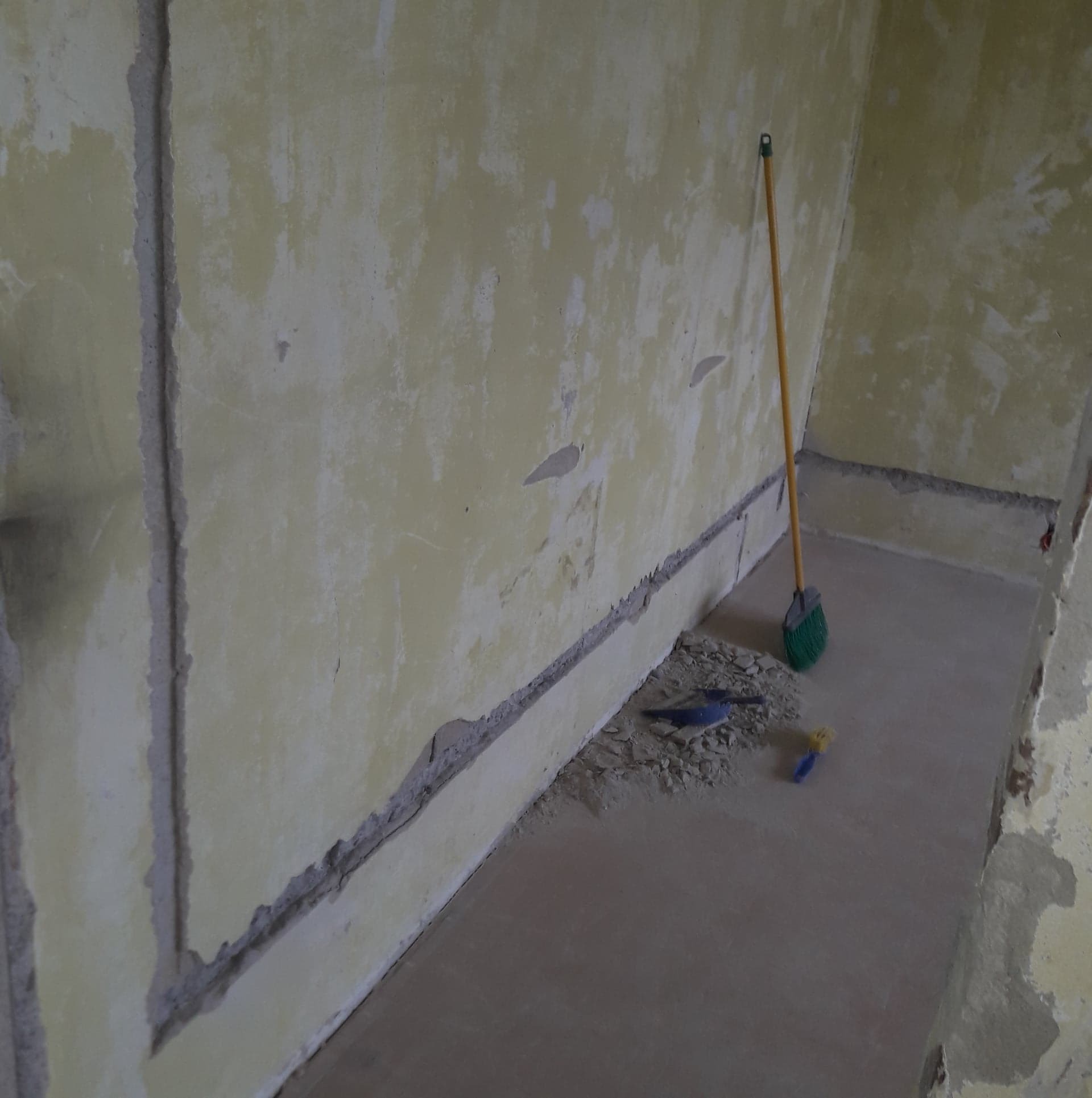 Przygotowanie do remontu: Odkurzanie podłogi po skuciu tynku, widoczna stara, żółta ściana z resztkami farby, nowa wylewka na podłodze i narzędzia.