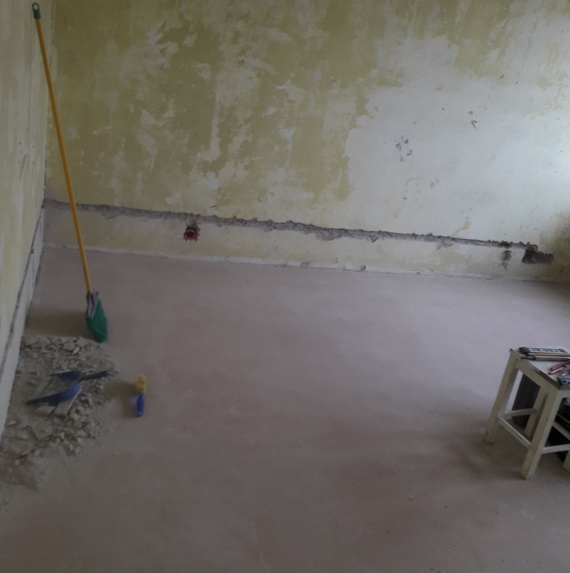 Świeżo wylana wylewka podłogowa w pomieszczeniu z resztkami starej farby na ścianach, widoczne narzędzia: szpachelka, wiadro i drabinka.