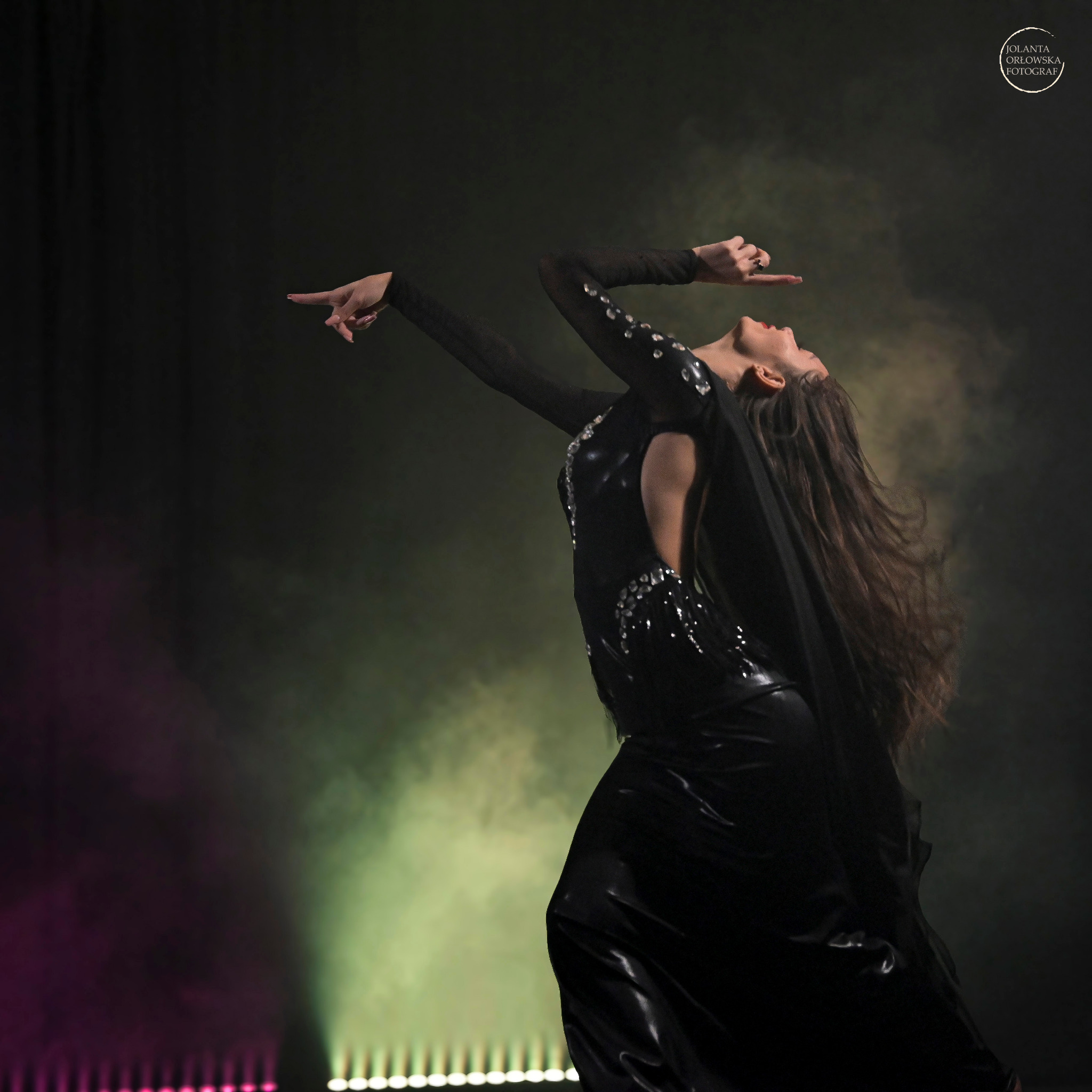 Kobieta w czarnej, błyszczącej sukni tanecznej, z głową odchyloną do tyłu i rękami wyciągniętymi w geście, na tle dymu i świateł scenicznych.