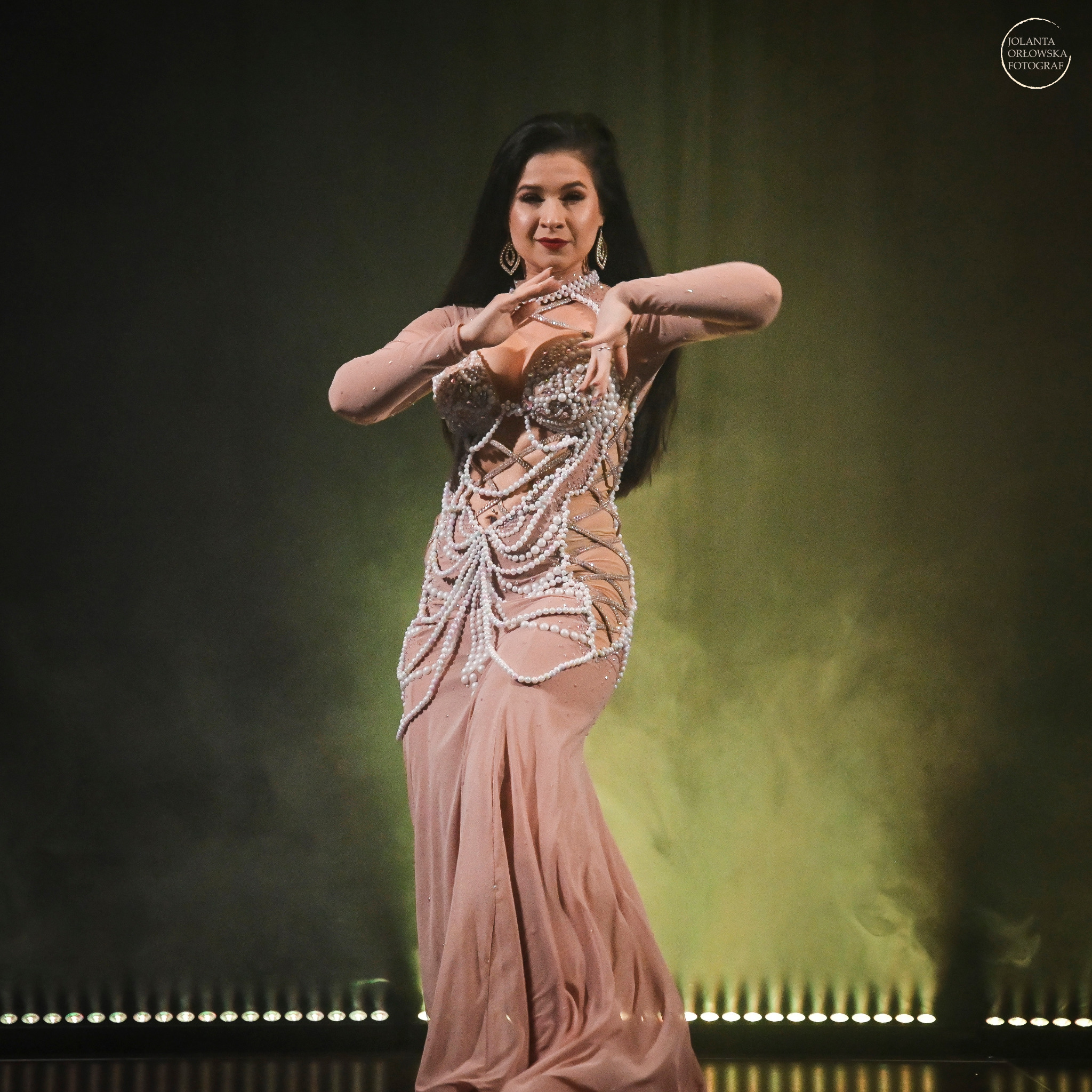 Tancerka w długiej, beżowej sukni zdobionej perłami, prezentująca układ taneczny na scenie z oświetleniem punktowym i zielonym dymem w tle.