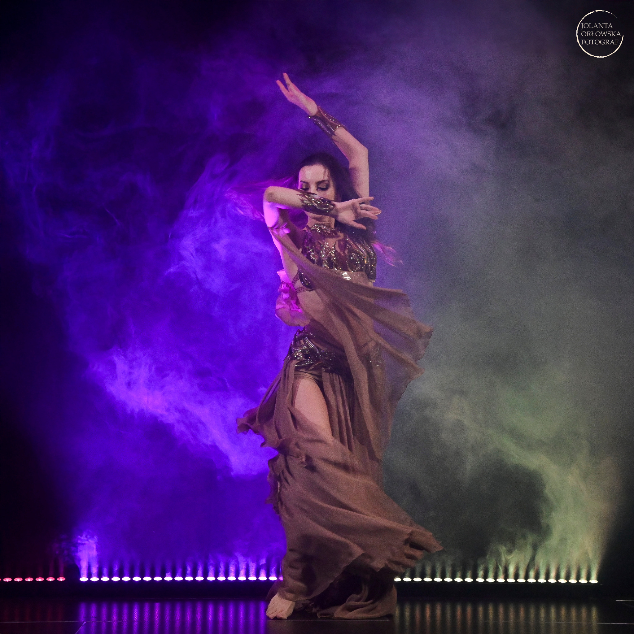 Tancerka w kostiumie z frędzlami, w ruchu, oświetlona fioletowym i zielonym światłem scenicznym, z efektem dymu.