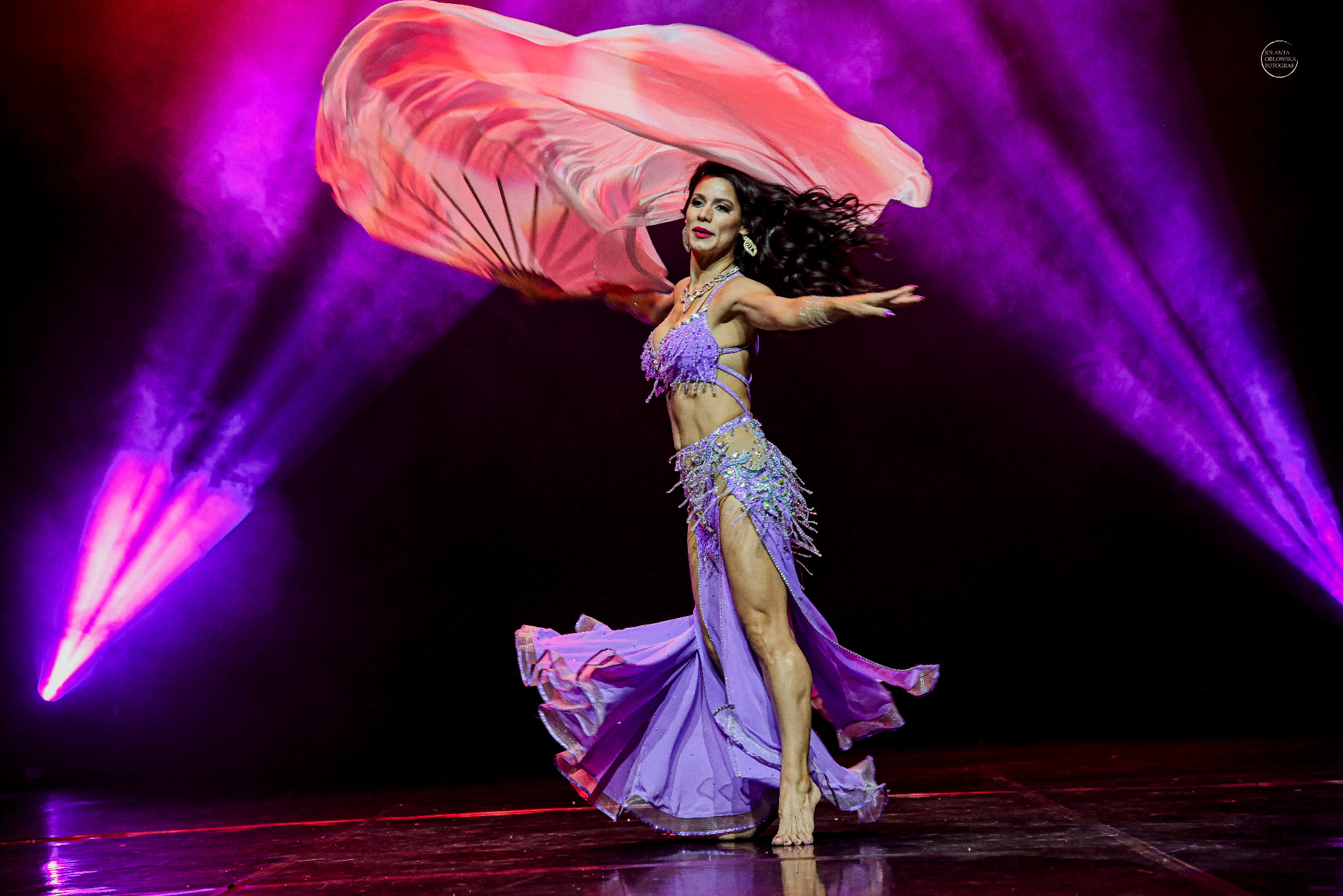 Kobieta w fioletowym, zdobionym stroju tanecznym, z rozpostartym, półprzezroczystym materiałem w dłoniach, na scenie oświetlonej fioletowymi reflektorami.