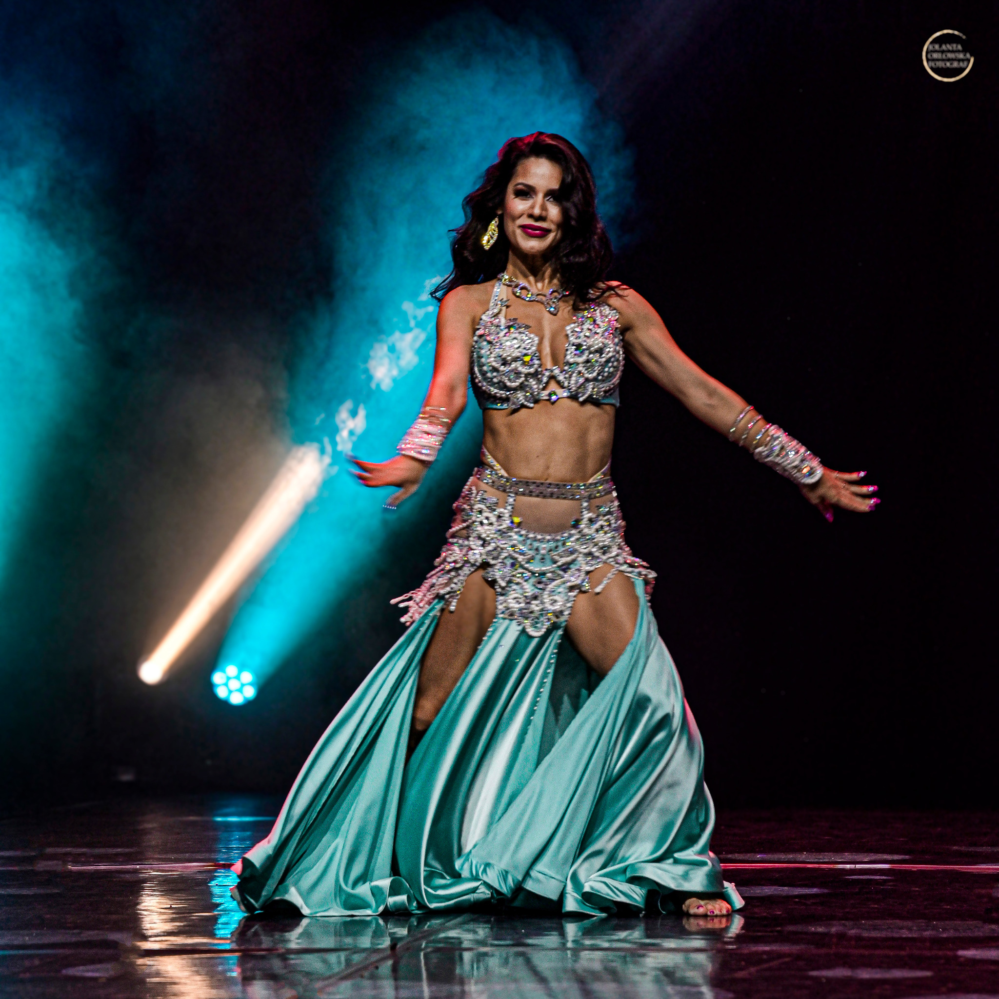 Uśmiechnięta kobieta w bogato zdobionym, turkusowym stroju tanecznym, z rozłożonymi rękami, na scenie z reflektorami i turkusową poświatą.