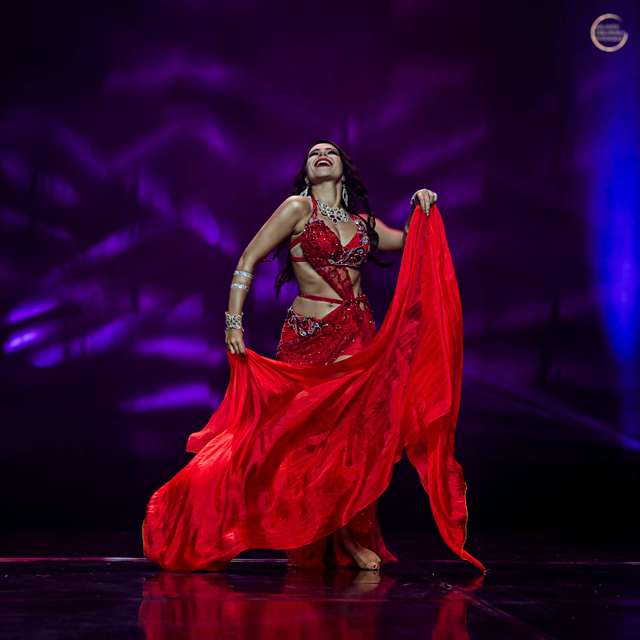 Uśmiechnięta tancerka w czerwonym, zdobionym stroju i z czerwonym szalem na scenie z fioletowym oświetleniem.