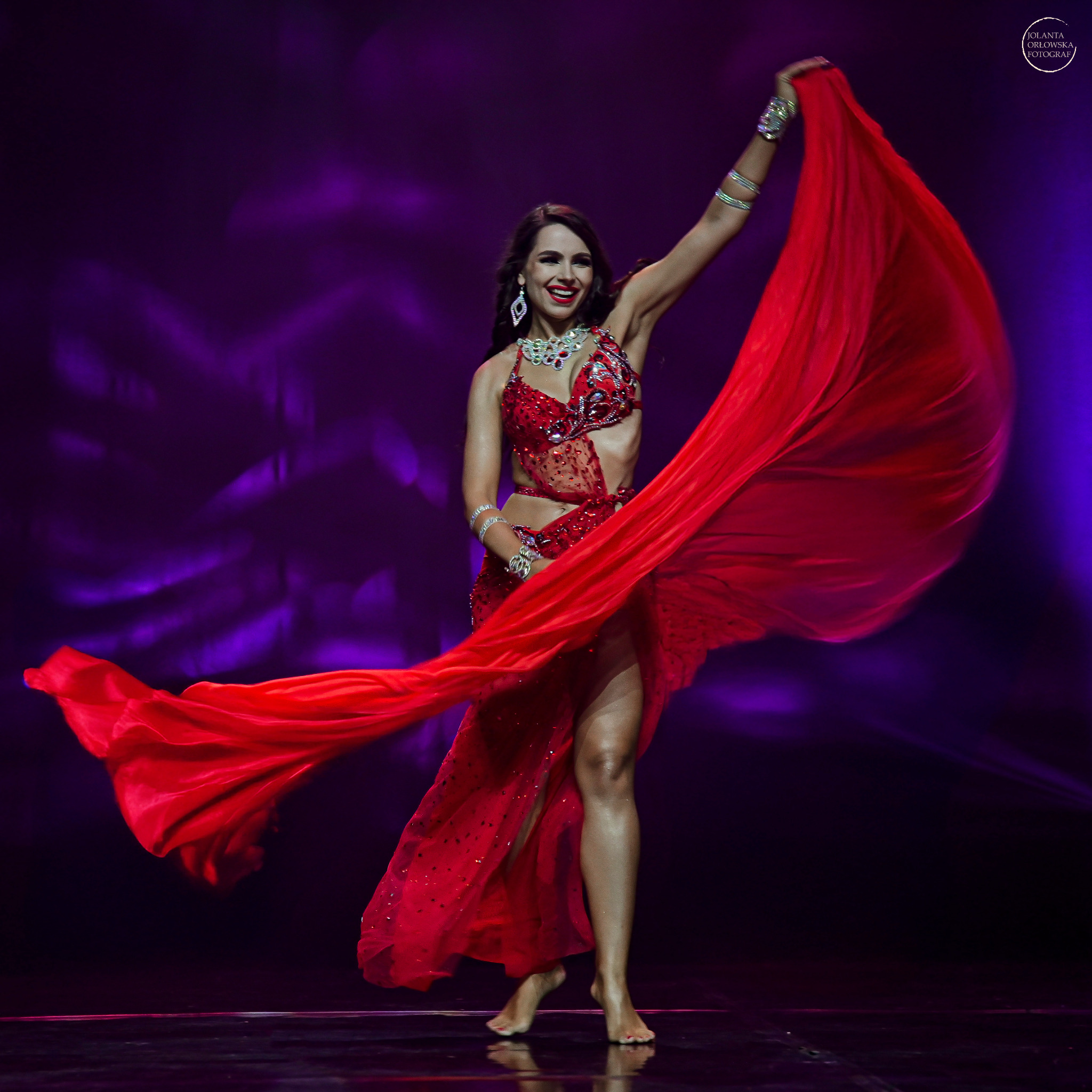 Uśmiechnięta kobieta w czerwonym, cekinowym stroju tanecznym, z rozpostartą czerwoną tkaniną w ruchu, występuje na scenie z fioletowym tłem w Myślenicach.