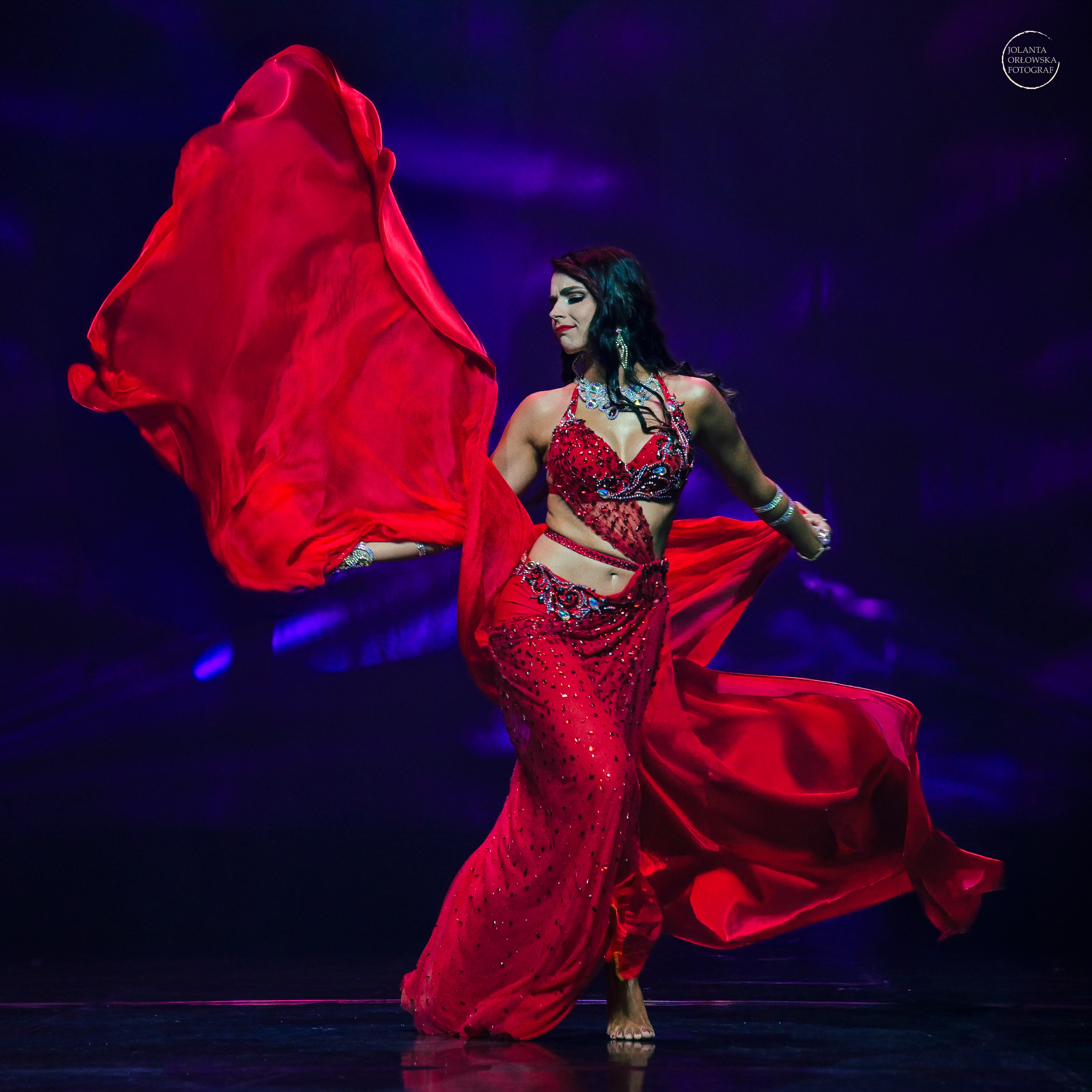 Kobieta w czerwonym, zdobionym kostiumie tanecznym z powiewającą tkaniną na scenie w niebieskim oświetleniu.