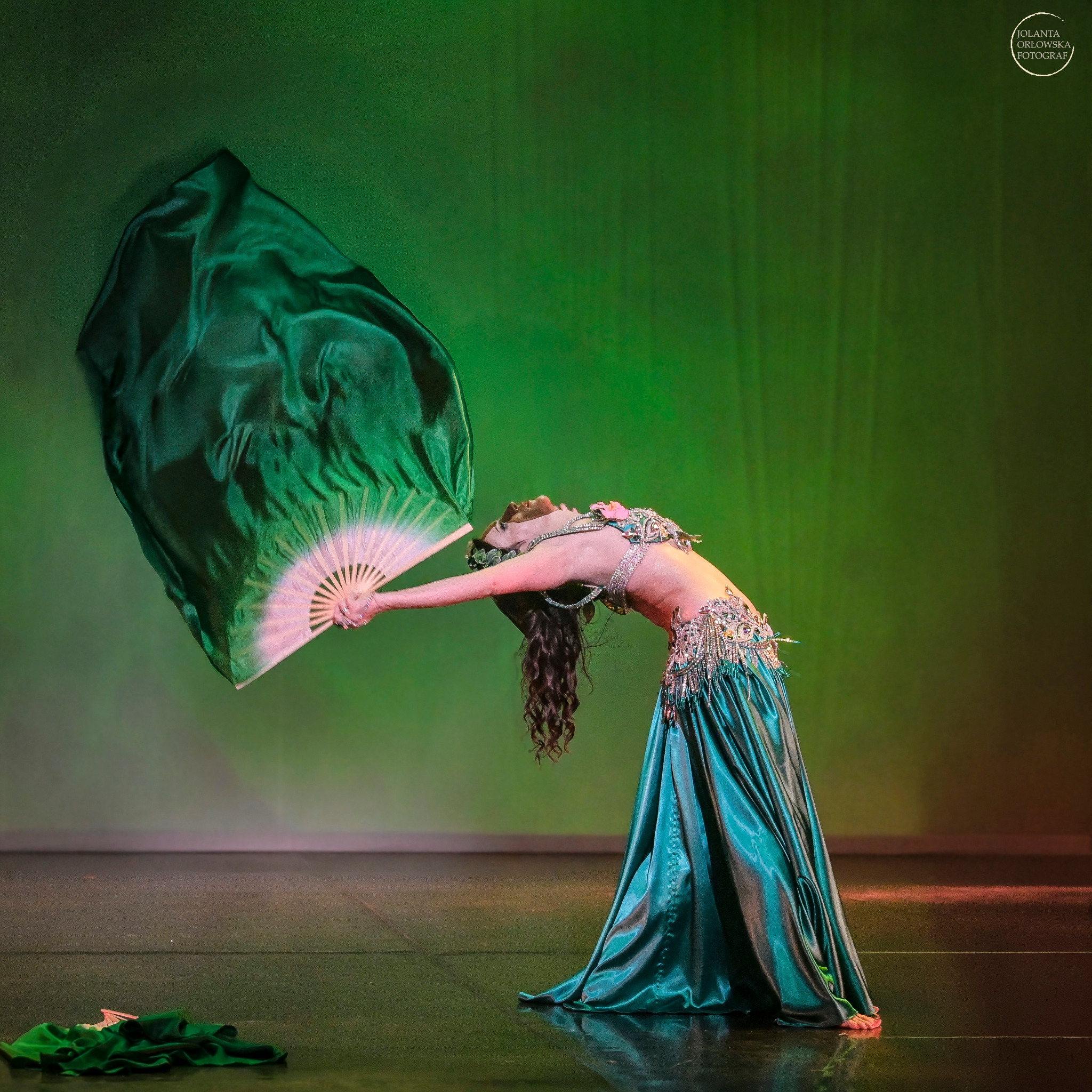 Tancerka w kostiumie z zielonego jedwabiu, wygina się do tyłu trzymając wachlarz i zwiewny materiał, oświetlona zielonym światłem scenicznym.
