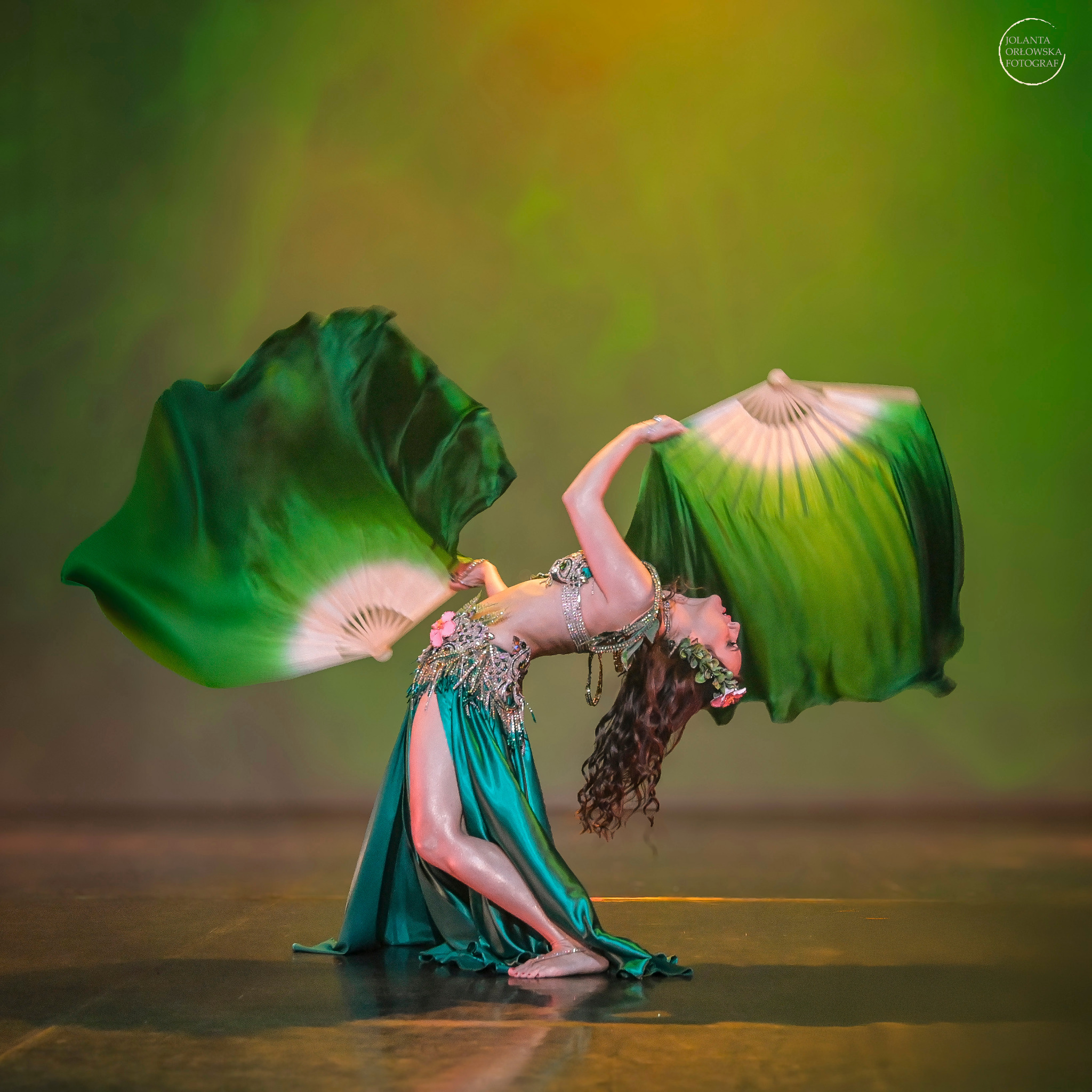 Tancerka w zielonym kostiumie, z wachlarzami z zielonego jedwabiu z białymi końcówkami, w wygiętej pozie tanecznej na scenie, z logo fotografa w prawym górnym rogu.