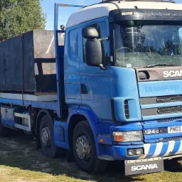 Niebieska ciężarówka Scania z pomarańczowym żurawiem i ładunkiem w czarnej skrzyni na tle zieleni i niebieskich zbiorników.