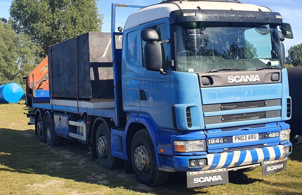 Niebieska ciężarówka Scania z pomarańczowym żurawiem i ładunkiem w czarnej skrzyni na tle zieleni i niebieskich zbiorników.