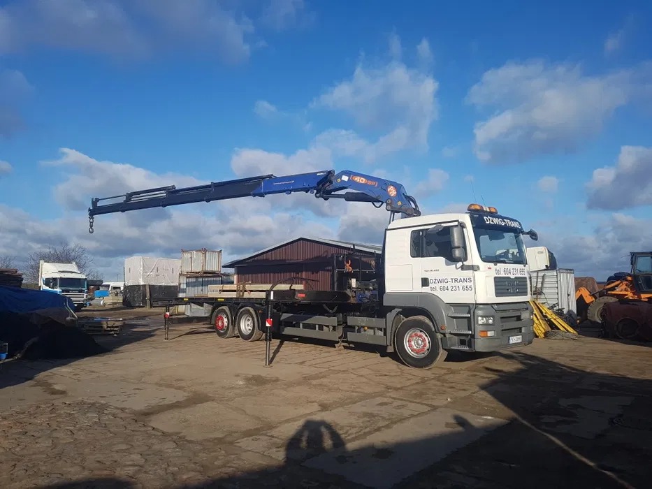 Ciężarówka z dźwigiem hydraulicznym marki MAN, zaparkowana na placu budowy, z wysuniętym ramieniem dźwigu i ładunkiem na platformie. Widoczne budynki w tle.