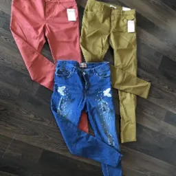 Trzy pary spodni: ceglaste, khaki i podarte jeansy z aplikacjami motyli z perełek, leżące na ciemnej podłodze z paneli.