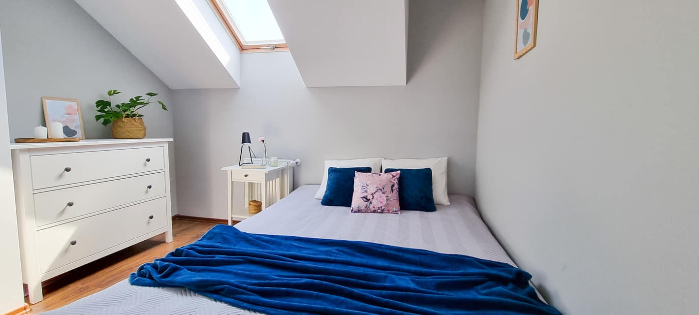 Pokój na poddaszu z łóżkiem, białą komodą i stolikiem nocnym, doświetlony oknem dachowym, z dekoracjami w postaci roślin, obrazków i poduszek.