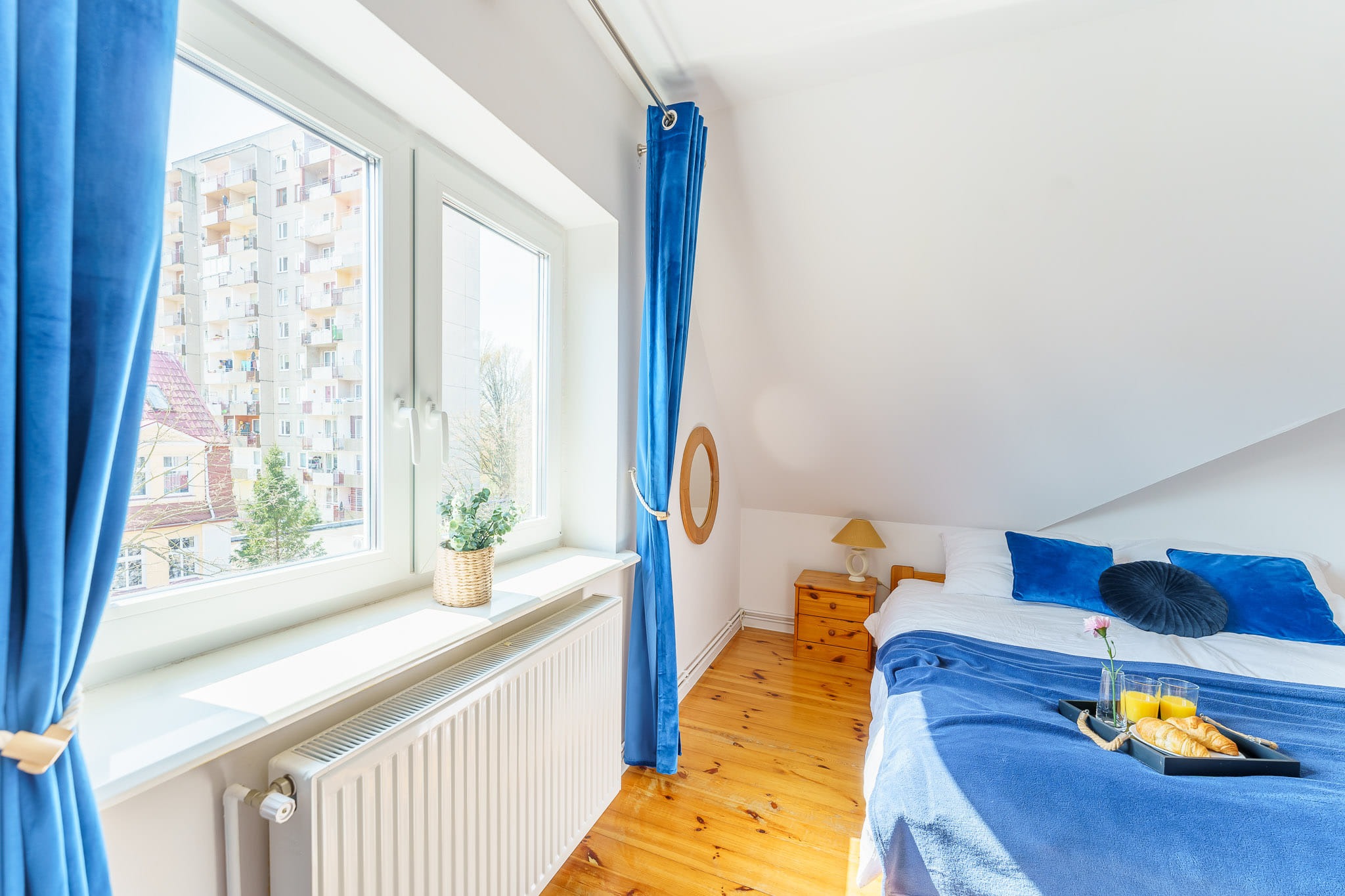 Jasna sypialnia na poddaszu z drewnianą podłogą, niebieskimi zasłonami i śniadaniem na łóżku: rogaliki, sok pomarańczowy i kwiat w wazonie.