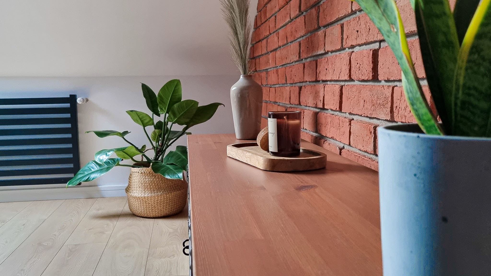 Fragment pokoju z roślinami w doniczkach, świecą na drewnianej tacy, wazonem z suszonymi trawami i dekoracyjną ścianą z cegły.