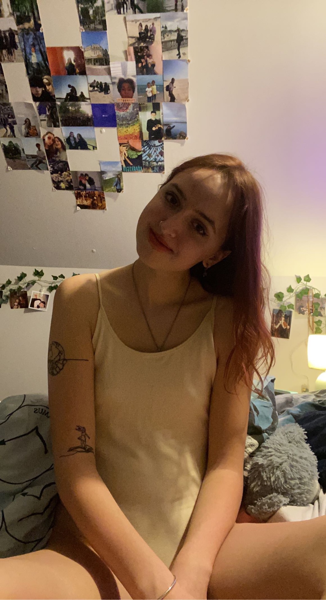 Portret młodej kobiety z rudymi włosami, z tatuażami na ramionach, siedzącej na łóżku w pokoju ozdobionym zdjęciami i girlandami.