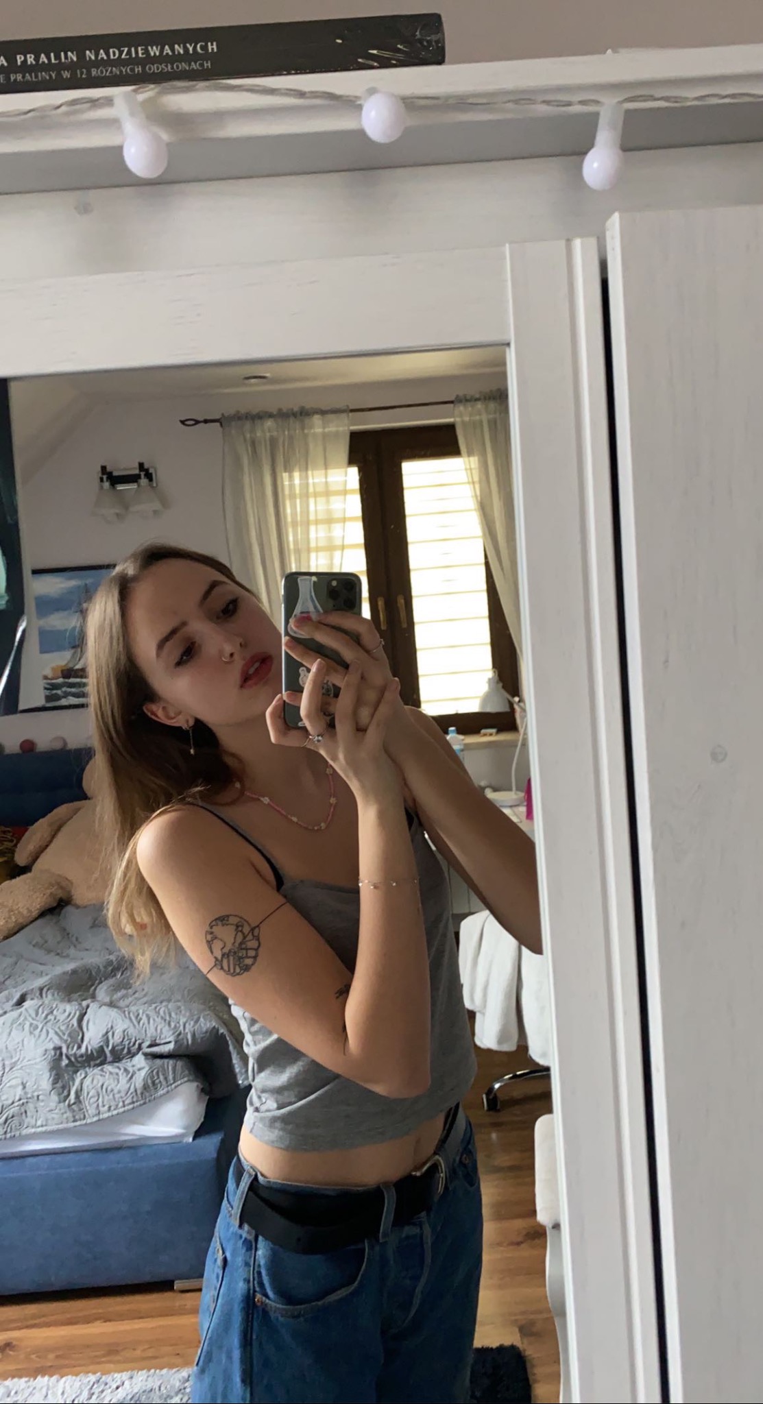 Młoda kobieta robi selfie w lustrze, widoczny tatuaż na ramieniu, ubrana w szary top i jeansy, w tle łóżko i okno.
