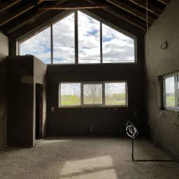 Surowe wnętrze domu w trakcie prac wykończeniowych, ściany pokryte szarą warstwą, widoczne okna z widokiem na zewnątrz, odsłonięta konstrukcja dachu, wystające przewody elektryczne.